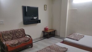 A room with a sofa, a small table, a TV mounted on the wall, and a bed. - Giriraj kripa (Vrindavan)