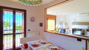 A dining area with a table set for four, a kitchen visible through an open doorway, and a large window with a view of greenery. - House in Luino with Green Garden and City Views (Luino)