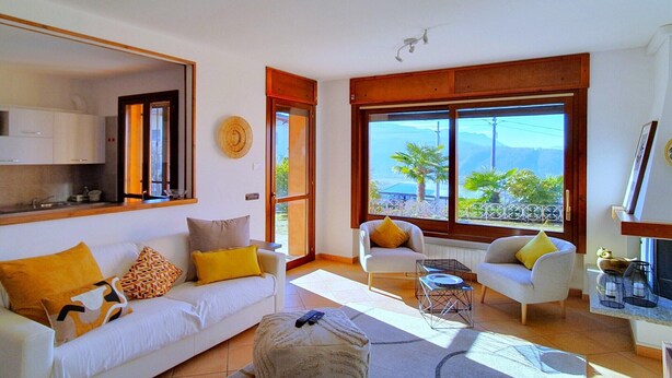 A living room with a white sofa, a small armchair, and a coffee table. There is a large window with a view of palm trees and mountains. - House in Luino with Green Garden and City Views (Luino)