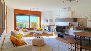 A modern living room with a sofa, ottoman, and coffee table, a dining area with a table and chairs, a fireplace, and a large window with a view. - House in Luino with Green Garden and City Views (Luino)