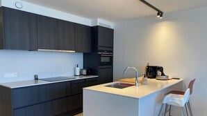 A modern kitchen with dark cabinetry, a central island with a sink, and a built-in oven. - Apartment in Nieuwpoort near Marina (Nieuwpoort)