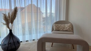 A room with a large window, a black vase with pampas grass, a beige sofa with a patterned pillow, and a wooden floor. - Apartment in Nieuwpoort near Marina (Nieuwpoort)