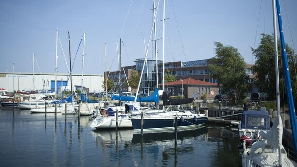 En småbåtshamn med flera segelbåtar förtöjda, en byggnad med ett rött tak och en klarblå himmel.