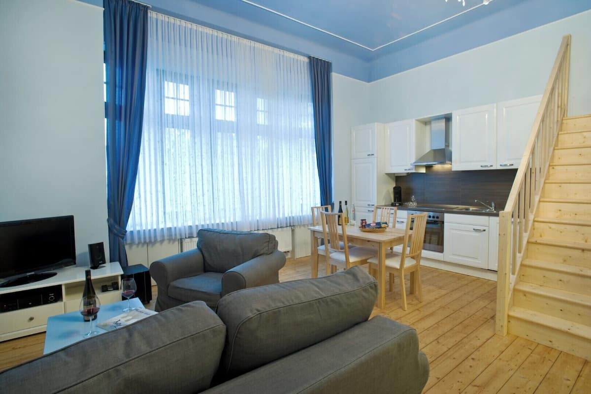 Una sala de estar moderna con un sofá gris, una mesa de comedor con sillas, un televisor y una zona de cocina con armarios blancos y una escalera de madera.