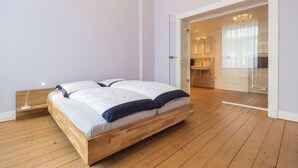 Une chambre avec un lit en bois, une table de chevet avec une lampe, et une vue sur la salle de bain et la cuisine à travers une porte ouverte.