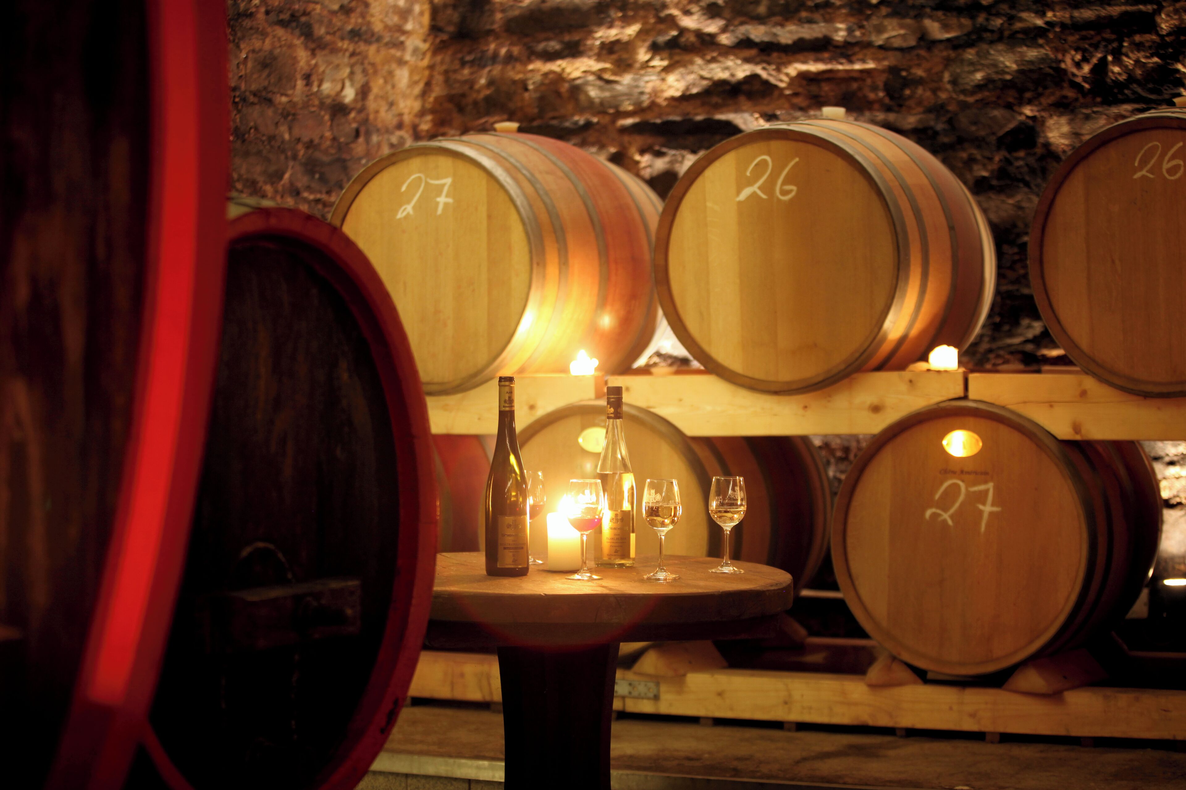 Barricas de madera con números, una botella de vino y dos copas sobre una mesa en una bodega.