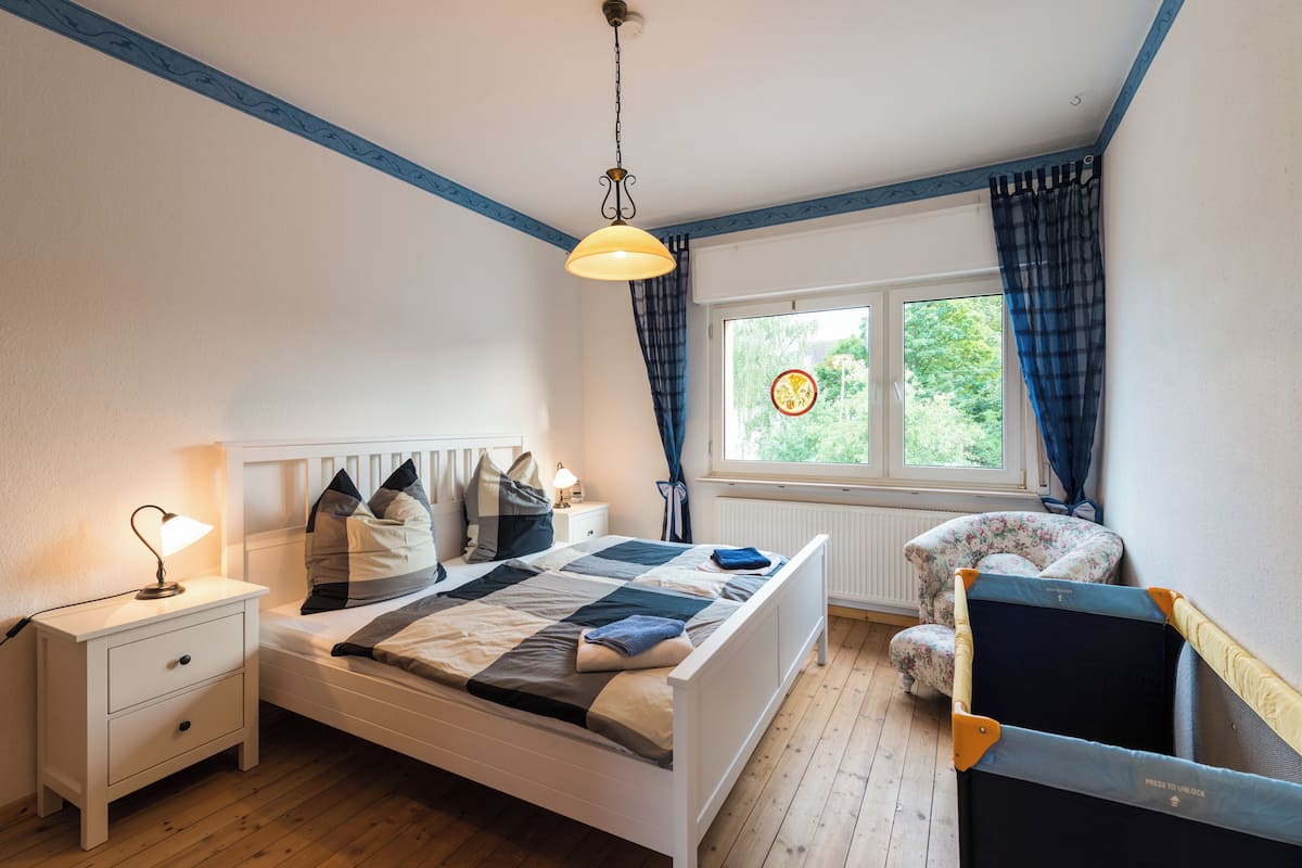 Un dormitorio con cama, mesita de noche, lámpara, ventana con cortinas y una silla.