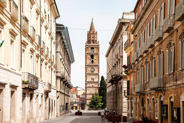 A historic street with a prominent clock tower in the center, flanked by classical buildings with balconies and arched windows.