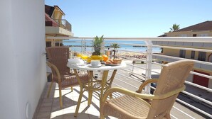 A balcony with wicker chairs and a table set with fruits and a cup, overlooking a beach with buildings in the background.