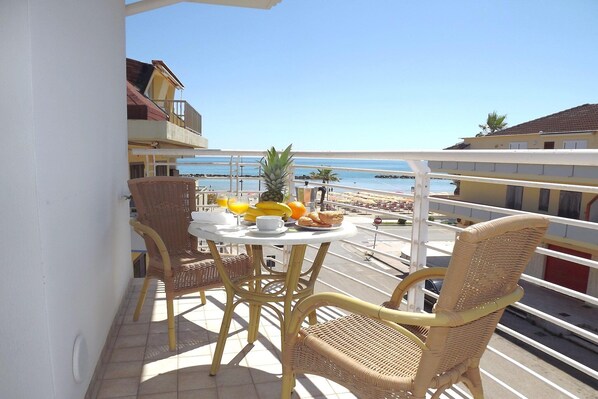 A balcony with wicker chairs and a table set with fruits and a cup, overlooking a beach with buildings in the background.