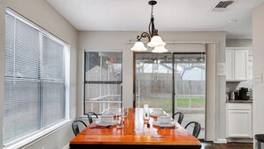 A dining room with a wooden table set for six, large windows with blinds, a kitchen area with white cabinets, and a hanging light fixture.