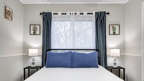 A neatly made bed with a white and blue bedspread, two matching pillows, and a black headboard. There are two bedside tables with lamps, and the room has a window with sheer white curtains and dark blue drapes.
