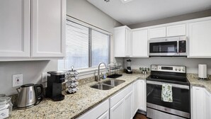 A kitchen with white cabinets, a stainless steel stove, a microwave, a coffee maker, and a window with blinds.