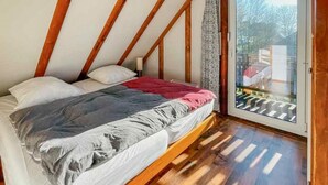 Un dormitorio con techo inclinado, vigas de madera, una cama con edredón gris y blanco, y vista a través de un ventanal.