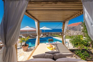 A covered outdoor seating area with a pool, lounge chairs, and a view of the sea.