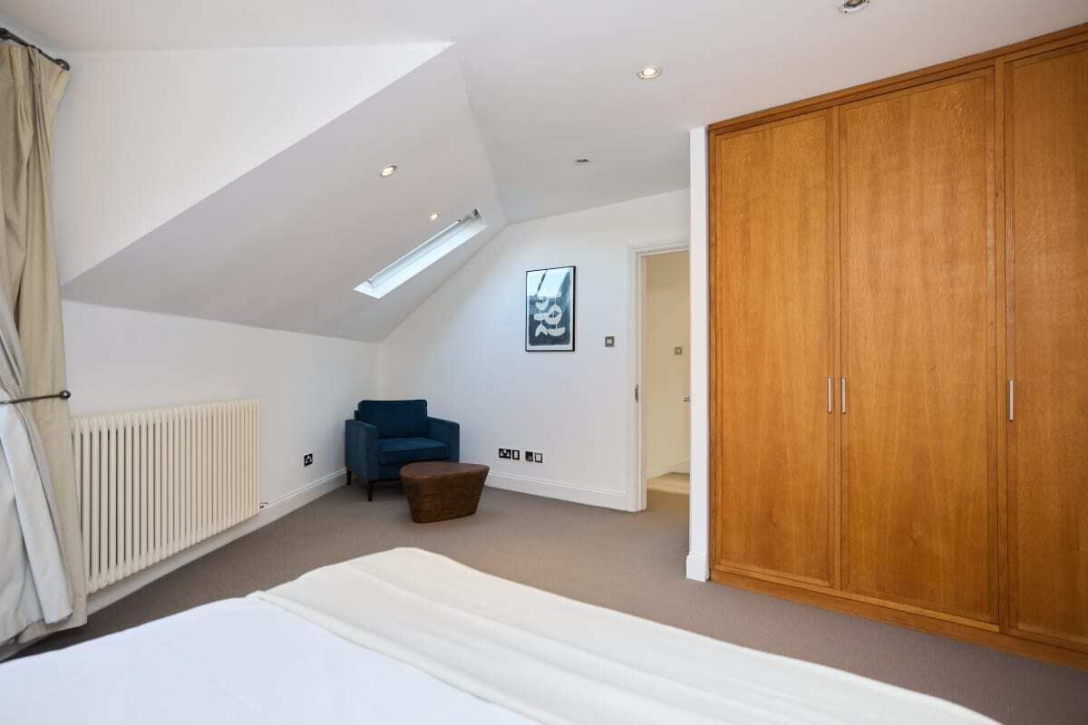 A bedroom with a sloped ceiling, a bed, a wardrobe, a chair, and a radiator.