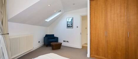 A bedroom with a sloped ceiling, a bed, a wardrobe, a chair, and a radiator.