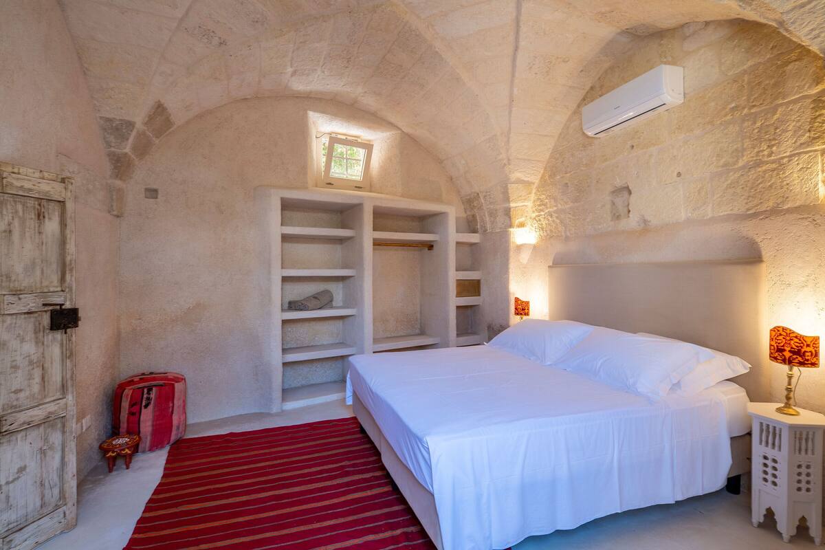 Ein Schlafzimmer mit Steindecke, einem Bett mit weißer Bettwäsche, einem roten Teppich, einem Kleiderschrank und einem kleinen Tisch mit Lampe.