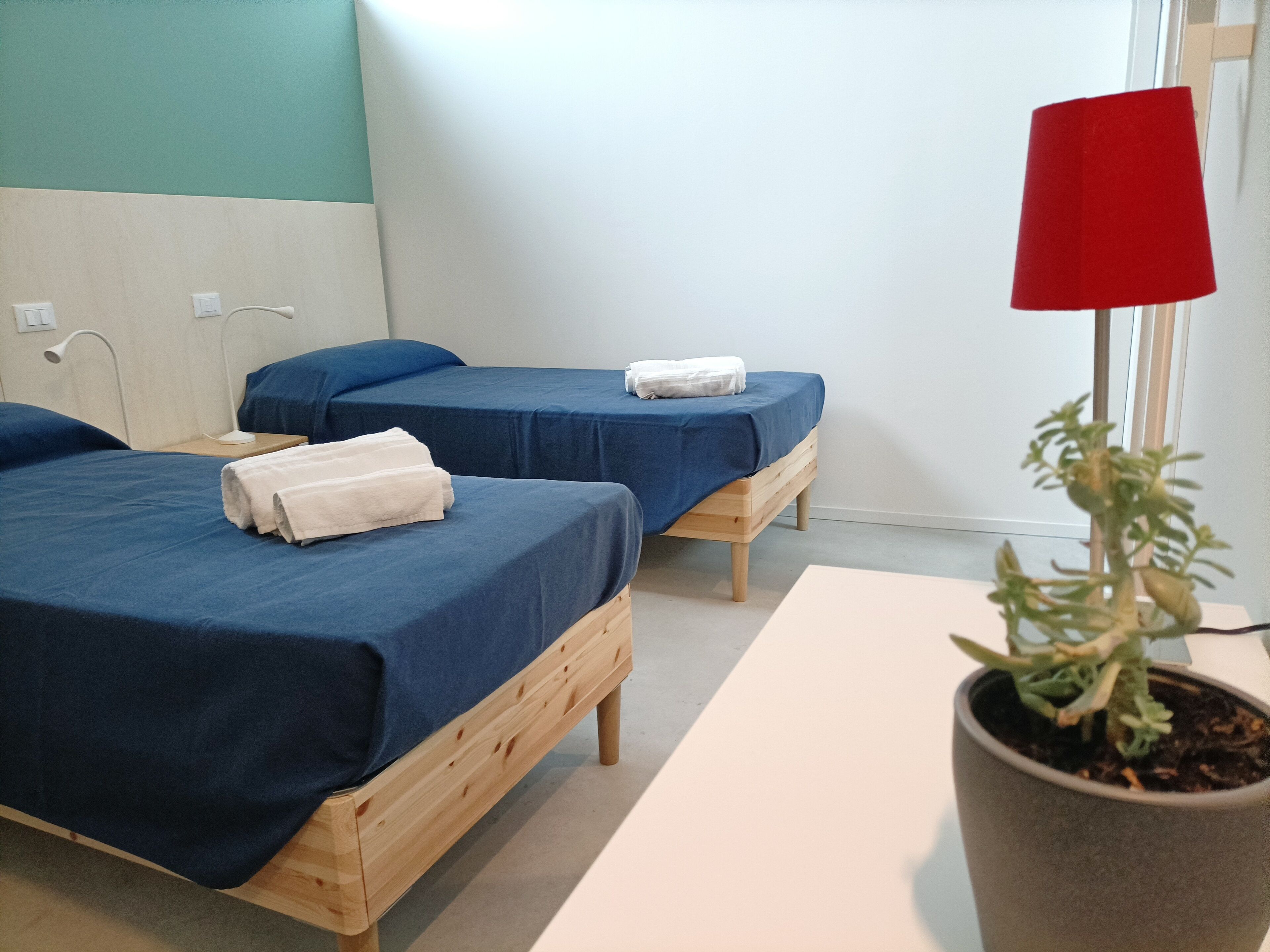 A room with two wooden beds, each with a blue bedspread and white towels. There is a red lamp on a table, and a potted plant on the floor.