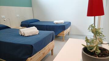 A room with two wooden beds, each with a blue bedspread and white towels. There is a red lamp on a table, and a potted plant on the floor.