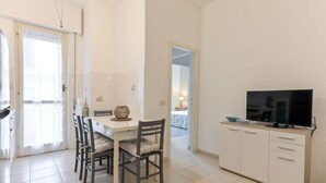 Un salon moderne et bien éclairé, comprenant un coin repas, un téléviseur et une cuisine visible à travers une porte ouverte.