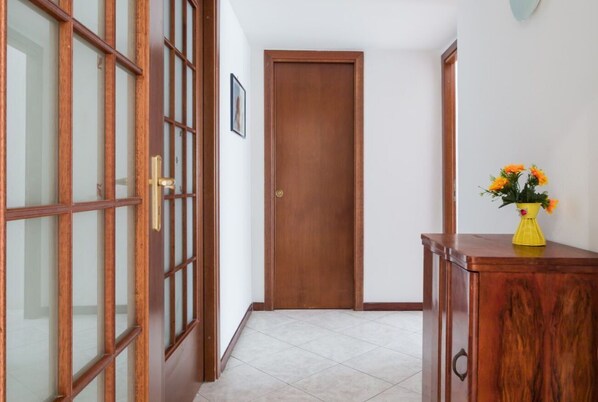 A hallway with a wooden door, a glass door, and a wooden cabinet with a vase of flowers. - Camera Augusto 42 By Dimorra (NAPOLI)