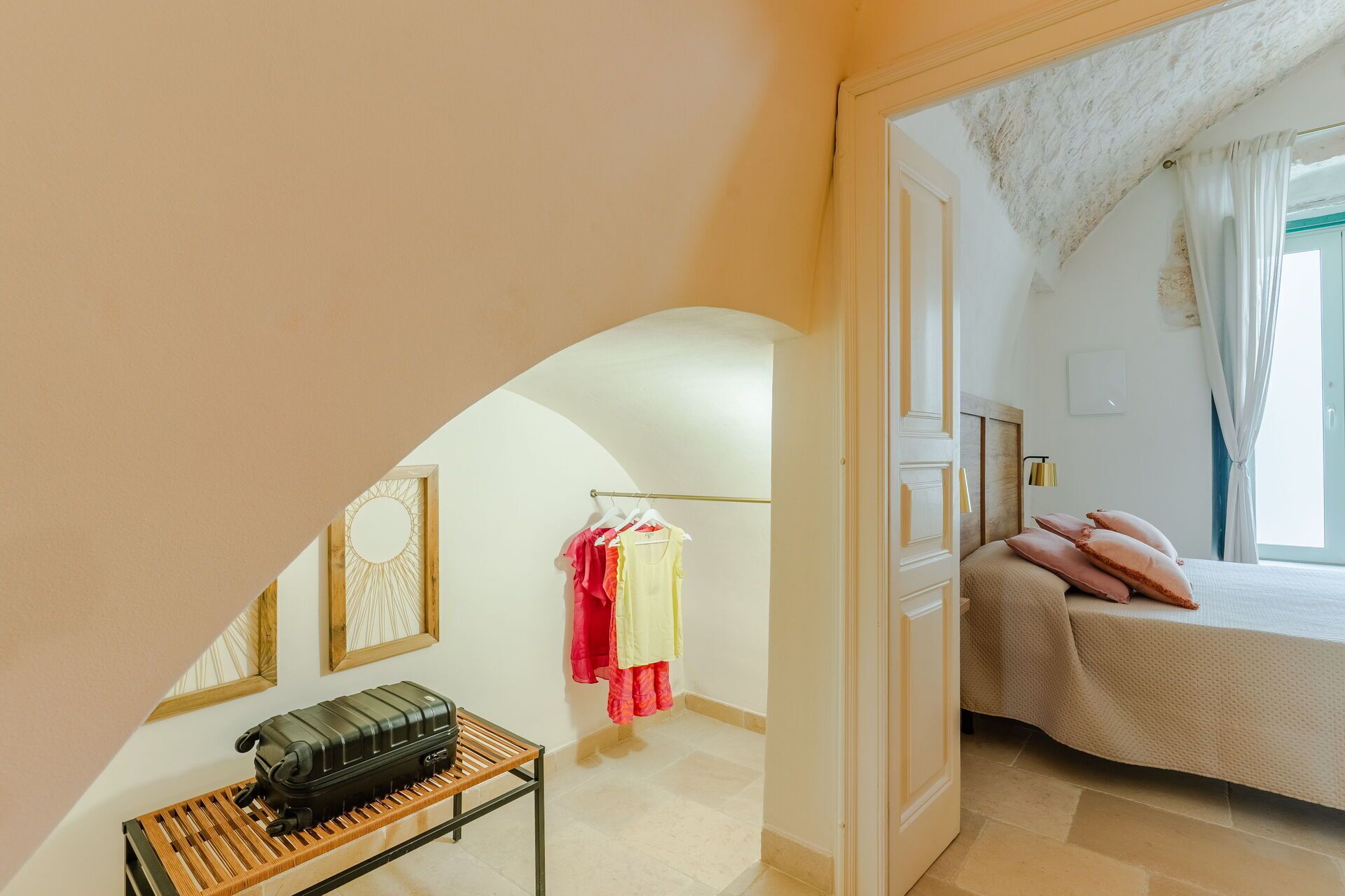 Quarto com cama, mala sobre uma mesa e um cabideiro com roupas vermelhas e amarelas.