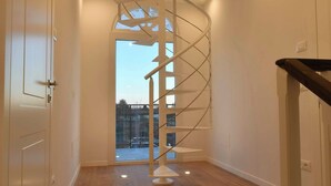 Ruime kamer met houten vloer, een witte deur en een trap met metalen leuning die naar een balkon leidt.