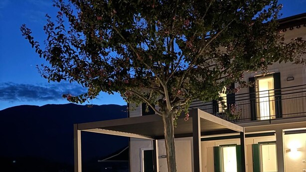 Een modern terras buiten met een pergola, een kleine boom met roze bloemen en een goed verlicht gebouw op de achtergrond.