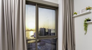 A room with a large window overlooking a cityscape and water, curtains, a white shelf with plants, and a wall-mounted picture frame. - Chic 2 BR in Vida Residences Canal View (Dubai)