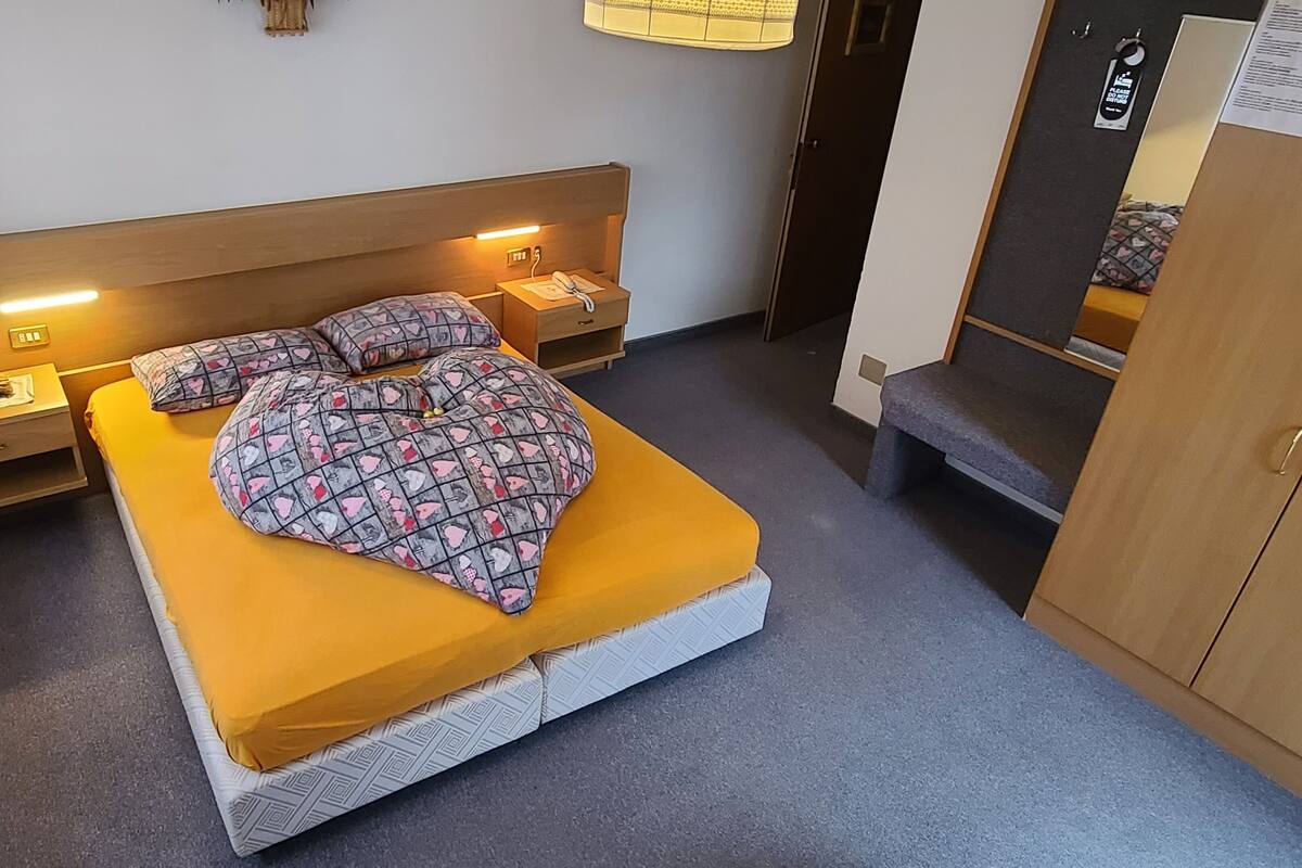 A single bed with a patterned blanket, a bedside table with a lamp, and a small drawer.