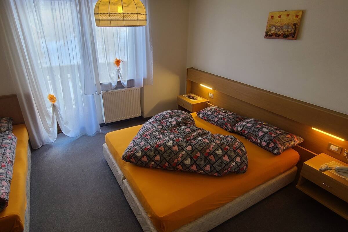 A hotel room with two single beds, a large bed, a nightstand, a lamp, a radiator, and a window with curtains.