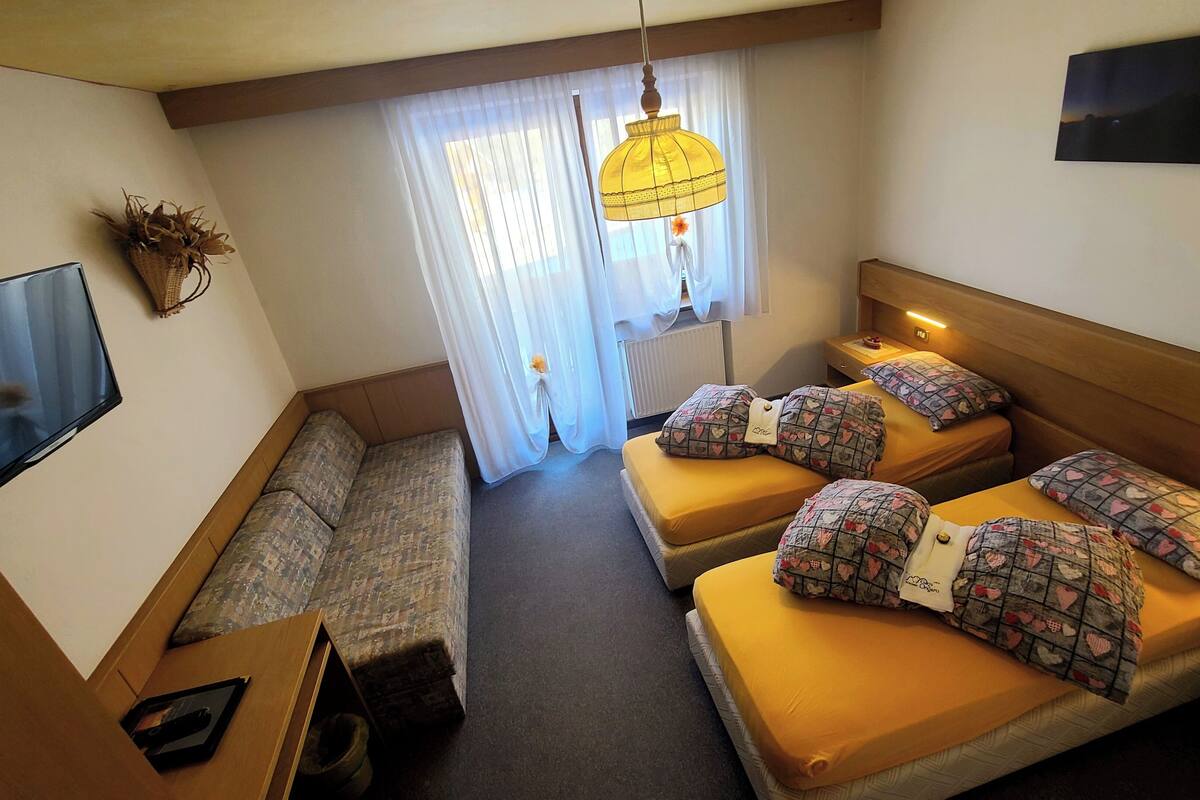 A compact hotel room with a sofa, two beds with patterned pillows, a TV, and a window with curtains.