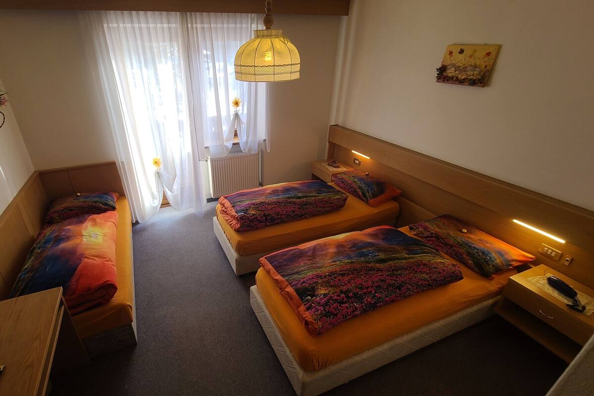 A hotel room with two beds, a wooden headboard, a bedside table, a lamp, a plant, and a window with curtains.