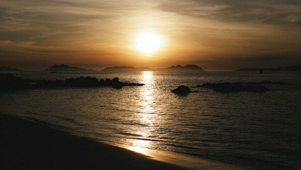 A sunset over a calm sea with rocks in the foreground and distant landforms.