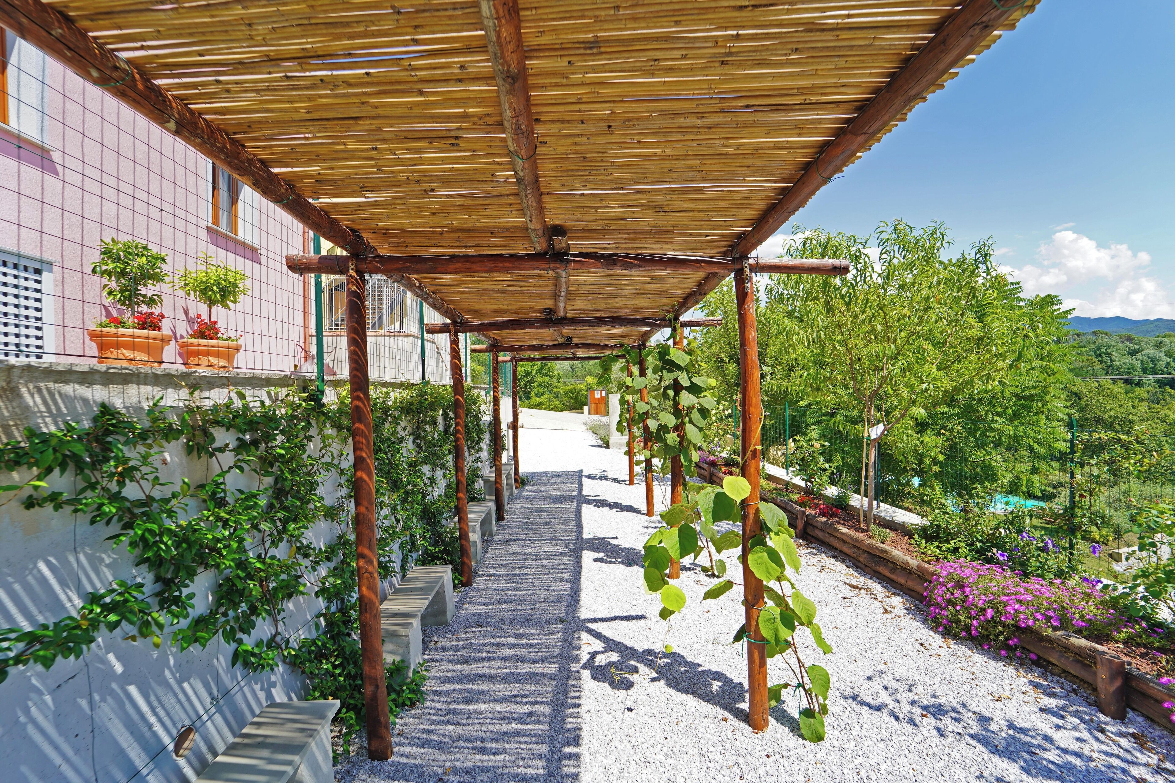 A covered walkway with a pergola structure, potted plants, and a gravel path leading to a building.