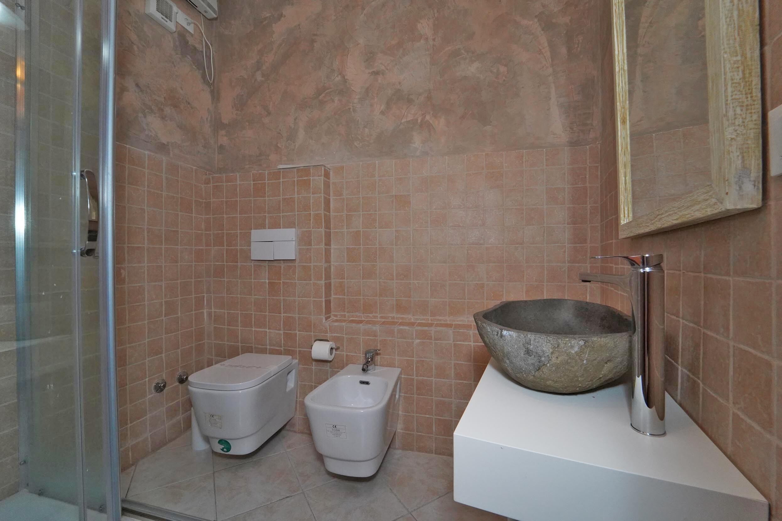 Un bagno con box doccia in vetro, un bidet, un water, un lavabo con bacino in pietra e un quadro appeso al muro.