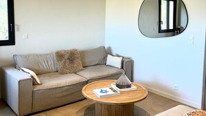 Una sala moderna con un sofá beige, una mesa redonda de madera y un reposapiés estampado. Un espejo grande está montado en la pared y una ventana proporciona luz natural.