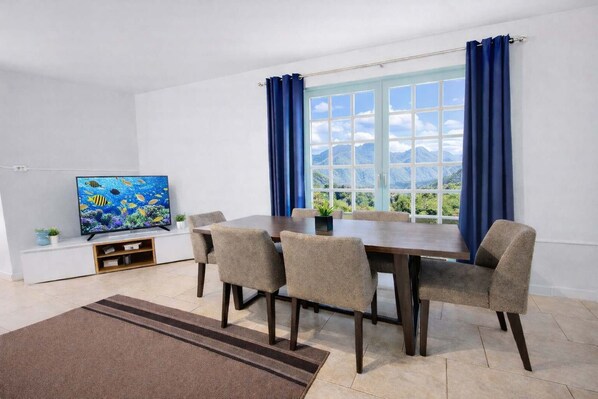 Una zona de comedor con una mesa y sillas, un televisor sobre un mueble bajo, y una ventana con cortinas.
