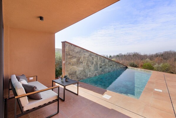 A modern outdoor pool area with a stone wall, a lounge chair, and a small table with a bottle and glass.