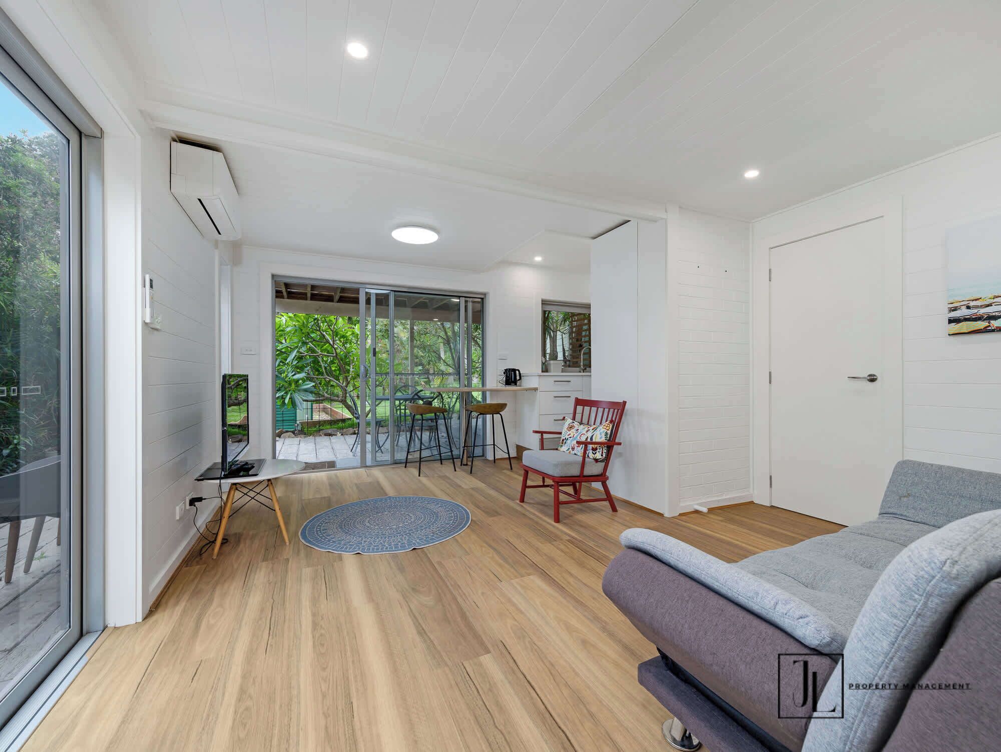 A modern living room with a sofa, a dining area with chairs, a kitchen with a sink and cabinets, and a sliding glass door leading to a garden.