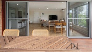 Un coin repas avec une table et des chaises en bois, un téléviseur en arrière-plan et une porte-fenêtre coulissante donnant sur un balcon.