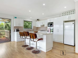 A modern kitchen with white cabinets, a central island with bar stools, a stainless steel refrigerator, and a window overlooking greenery.