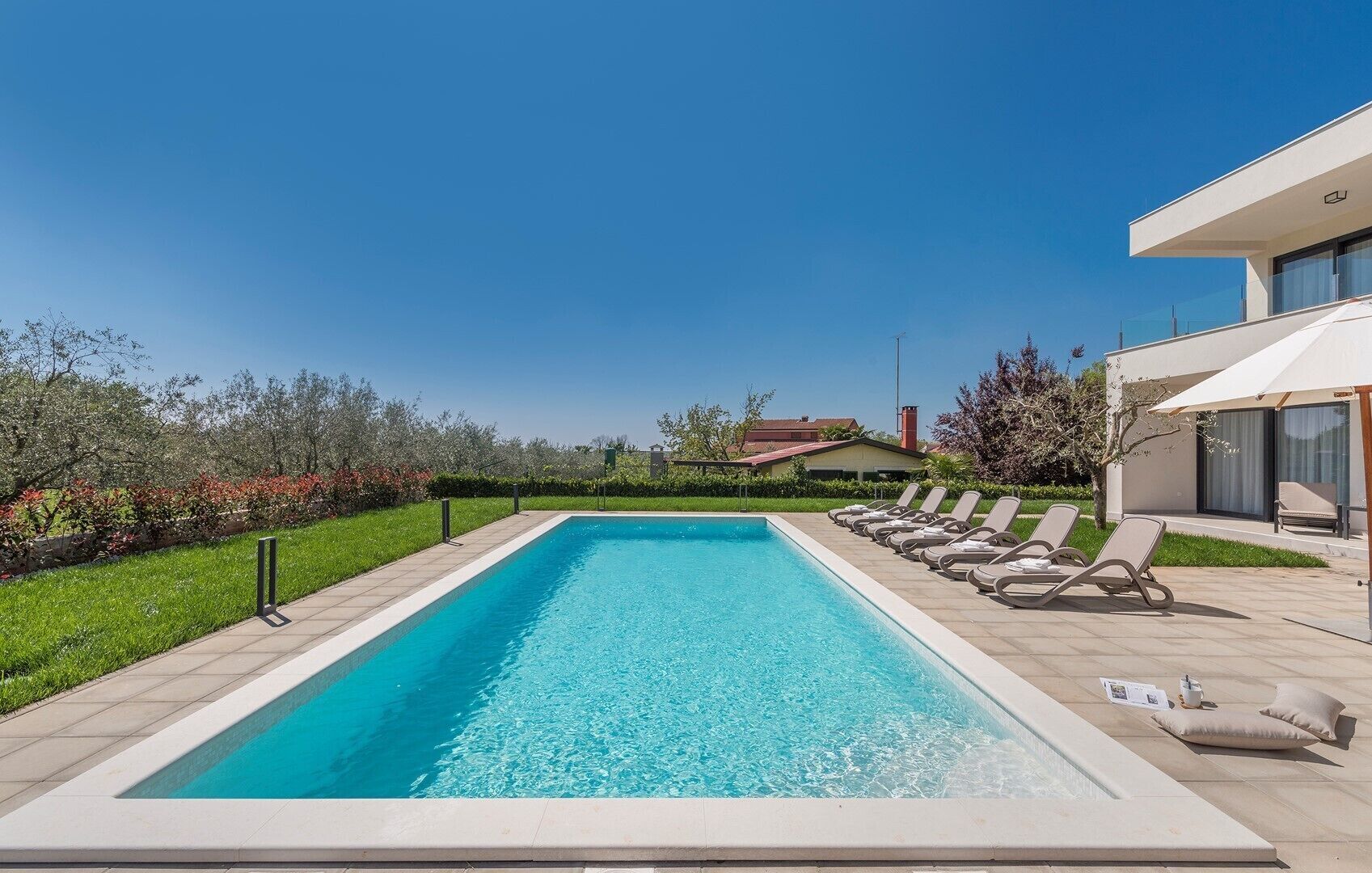 Una piscina rectangular con agua azul cristalina, rodeada por una terraza con baldosas. Hay sillas de descanso alineadas junto a la piscina, y un edificio moderno con grandes ventanales es visible al fondo.