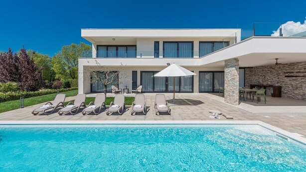 Ett modernt hus i två våningar med en swimmingpool, solstolar och en stenmur.