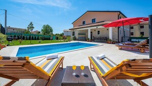 Piscina com espreguiçadeiras e uma mesa com bebidas, cercada por um edifício com um guarda-sol vermelho.