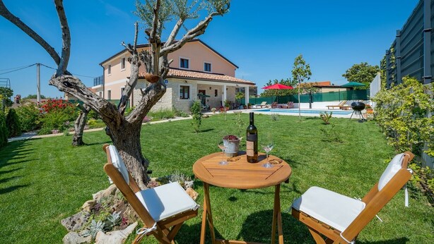 Um pátio com duas cadeiras de madeira e uma mesa, uma garrafa de vinho e uma piscina ao fundo.