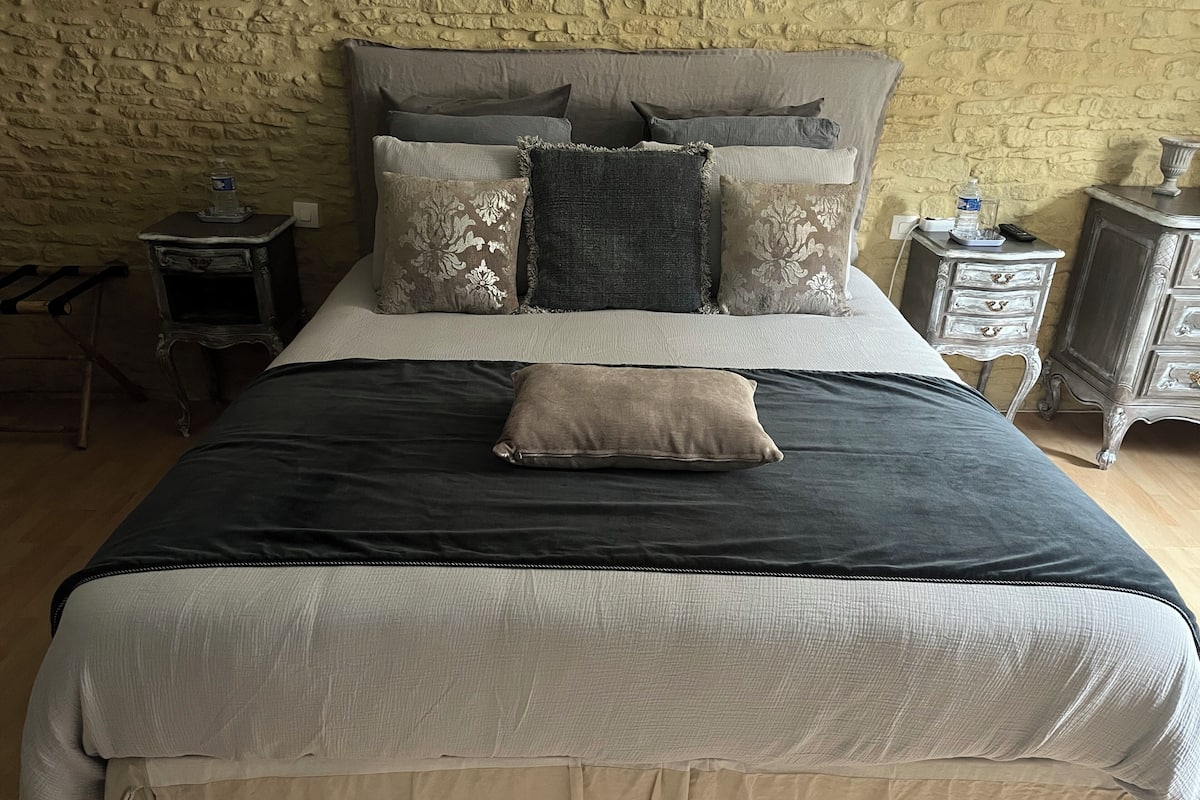 Un lit avec une tête de lit sombre, plusieurs oreillers, une table de chevet avec une bouteille d’eau et une petite table de nuit avec une lampe.