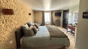 A bedroom with a stone wall, a bed with multiple pillows, a chair, a desk, a TV, and a sign indicating WiFi availability. - Chambres d'Hôtes de l'Ancienne Auberge (Thaon)