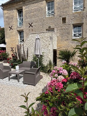 Un bâtiment en pierre doté d’un patio aménagé avec du mobilier d’extérieur et d’un jardin où poussent des fleurs roses.